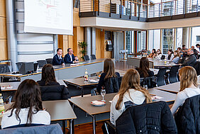 Bezirkstagspräsident Peter Daniel Forster und Bezirksrat Herbert Lindörfer sitzen vorne im Sitzungssaal an einem Tisch und befinden sich im Gespräch mit der Schulklasse, die im Sitzungssaal verteilt sitzt.