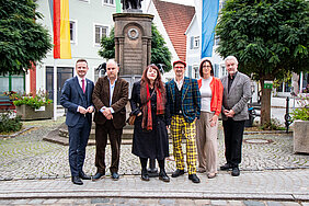 Bezirkstagspräsident Peter Daniel Forster, Preisträger Matthias Egersdörfer, die beiden Förderpreisträgerin Julie Batteux und David Saam, Kulturreferentin des Bezirks Dr. Annett Haberlah-Pohl und für das Sinfonieorchesters der Hochschule für Musik in Nürnberg dessen Leiter Prof. Guido Johannes Rumstadt stehen vor einem Brunnen in Wolframs-Eschenbach