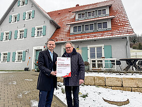 Bezirkstagspräsident Peter Daniel Forster überreicht in Unterwindsberg den Förderbescheid über 15.000 Euro. Die beiden Personen stehen vor dem sanierten Gebäude.