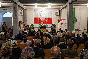 Foto von der Preisverleihung aus dem vergangenen Jahr. Bezirkstagspräsident Peter Daniel Forster steht am Rednerpult und begrüßt das Publikum, das auf Stühlen vor ihm sitzt.