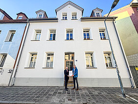 Für das Engagement in der Denkmalpflege überreicht Bezirkstagspräsident Peter Daniel Forster in Nürnberg den Förderbescheid. Die beiden Personen stehen vor dem sanierten Gebäude.