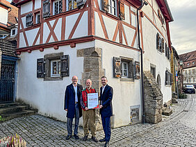 Bezirkstagspräsident Peter Daniel Forster überreicht den Förderbescheid im Beisein des Oberbürgermeisters von Rothenburg o.d.T. Dr. Markus Naser. Sie stehen vor dem sanierten Gebäude.