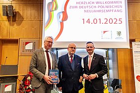 Prof. Dr. Peter Oliver Loew, Rafał Wolski und Bezirkstagspräsident Peter Daniel Forster stehen beim deutsch-polnischen Neujahrsempfang für ein Gruppenfoto vor der Leinwand im Sitzungssaal des Bezirksrathauses in Ansbach.